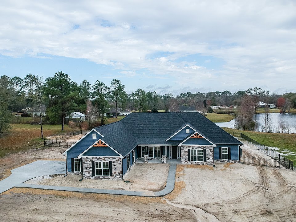 Two-story house with light siding, gabled roof, paved driveway, mature trees in the background, and blue sky with scattered clouds