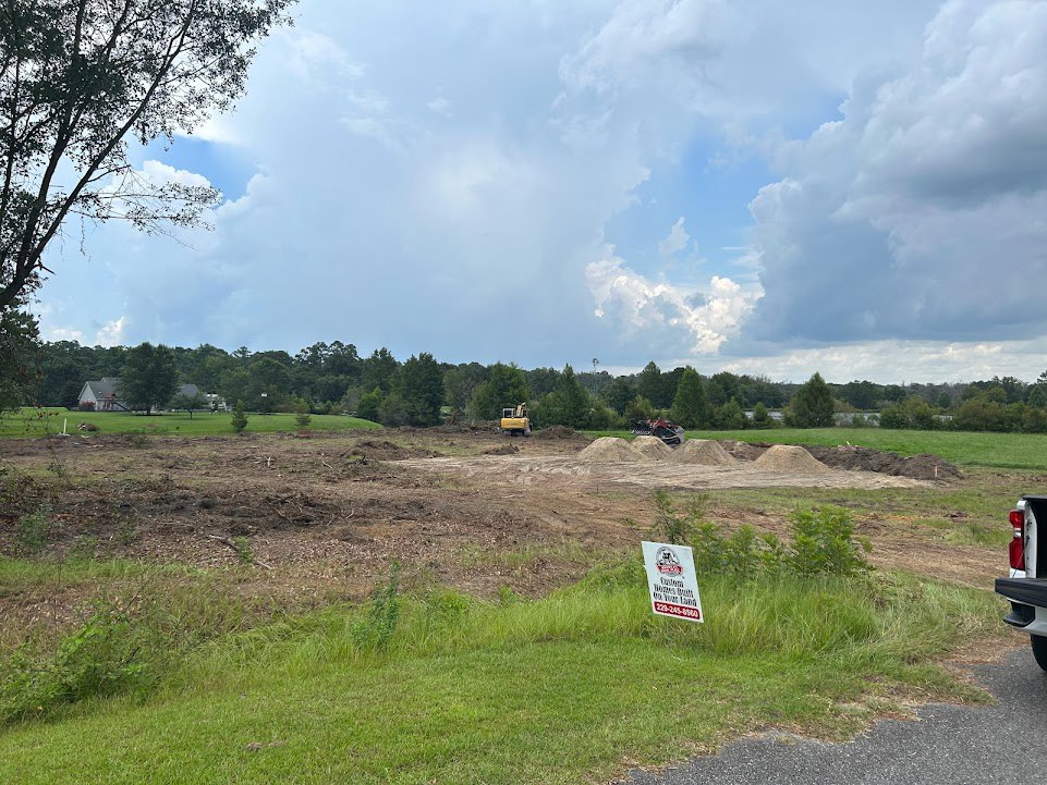 Grassy construction site with yellow tractor, white sign, and mature trees under partly cloudy blue sky