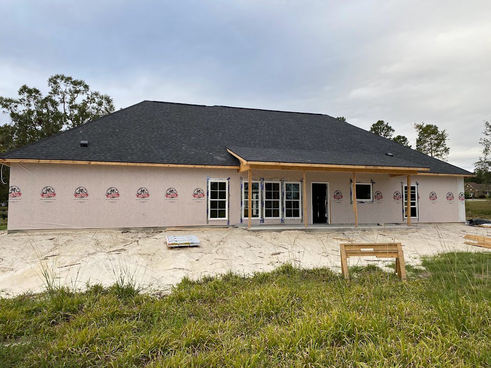 Partially built house with exposed framing, shingled roof, grassy yard, wooden bench, and scattered construction materials