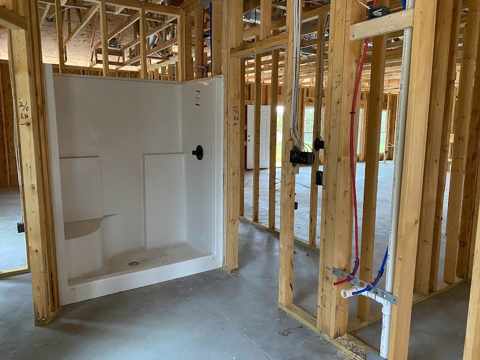 Bathroom under construction with exposed wooden ceiling beams, red electrical cable, unfinished shower with built-in shelf, debris scattered on plywood floor, partially insulated