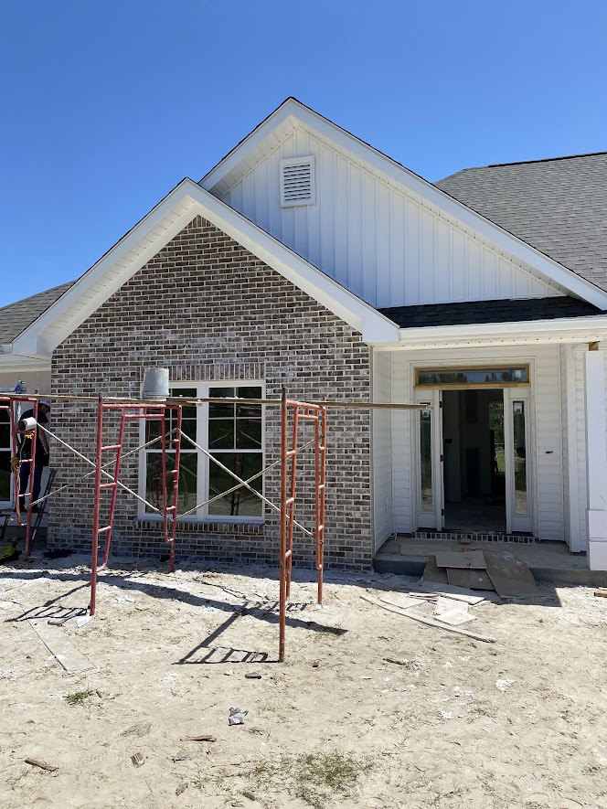 Red scaffolding set up against a partially constructed brick house with glass panel door and large window, surrounded by bare ground under a clear sky