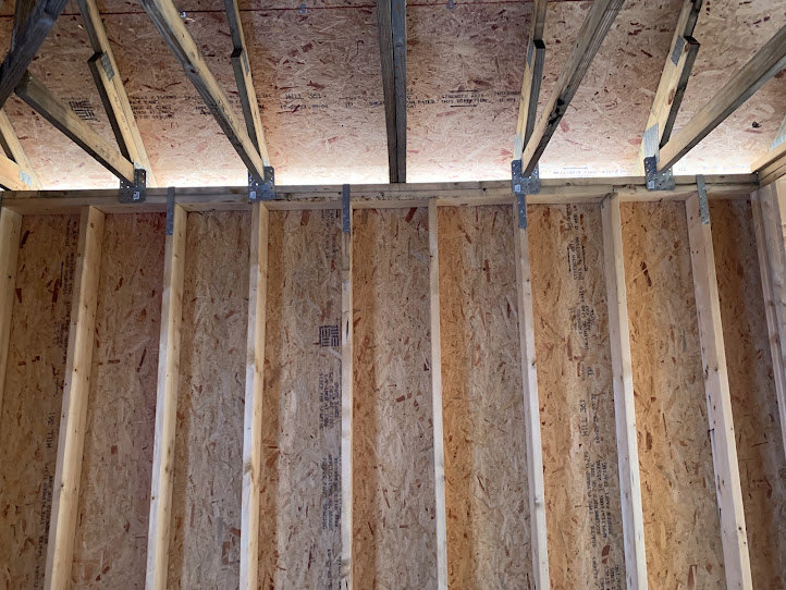 Exposed wood framing with metal brackets, unfinished beams and planks, close-up of a wood panel marked with black text, white pole with brown handle, construction materials and