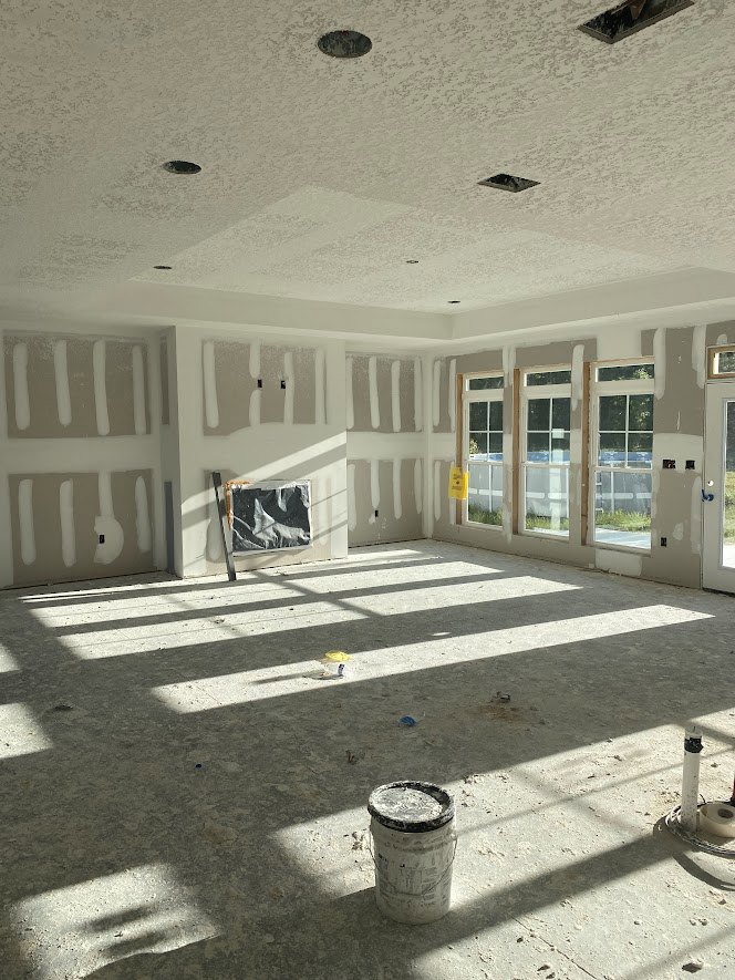 Bright room with white painted walls, large windows, a door, paint bucket with newspaper on top, and a piece of paper taped to the wall