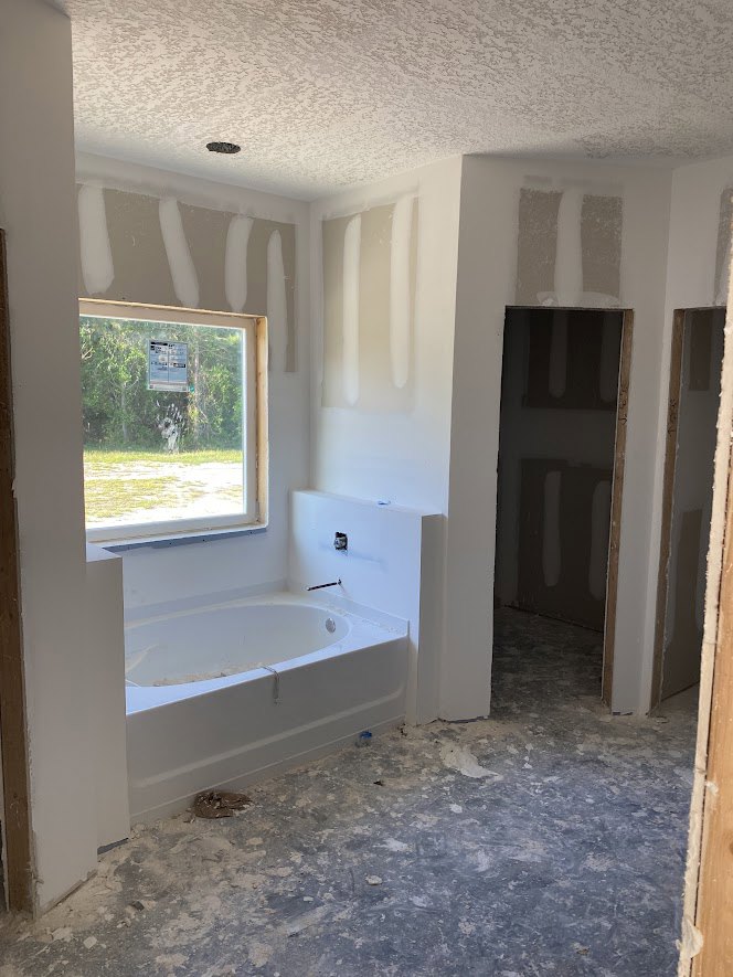 Bathroom with freestanding white tub beneath a window, unfinished plaster walls, dusty white floor, and partially installed door frame.