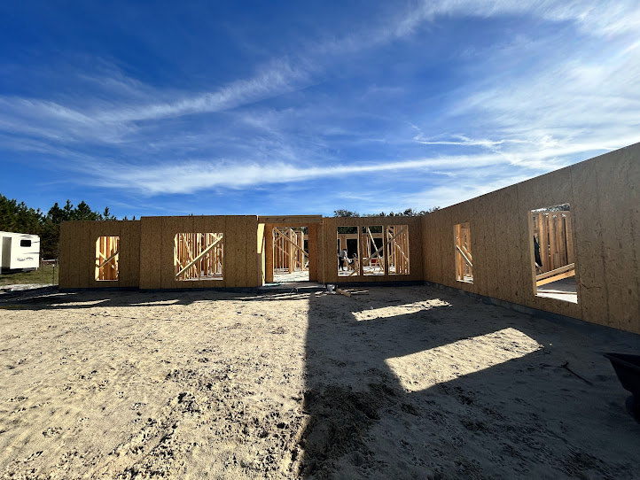 Framed custom home under construction with exposed wood, sandy ground, and clear blue sky