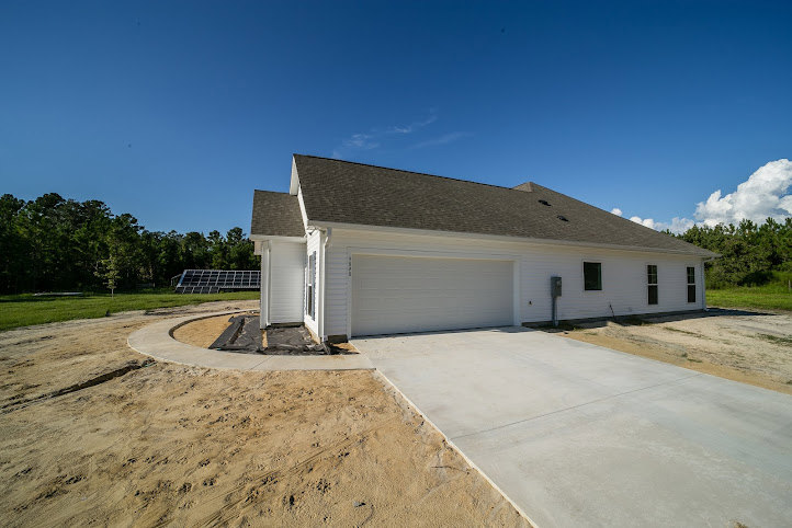 Modern two-story house with light siding, attached garage, paved driveway, rooftop solar panels, manicured lawn, and mature trees under a partly cloudy sky