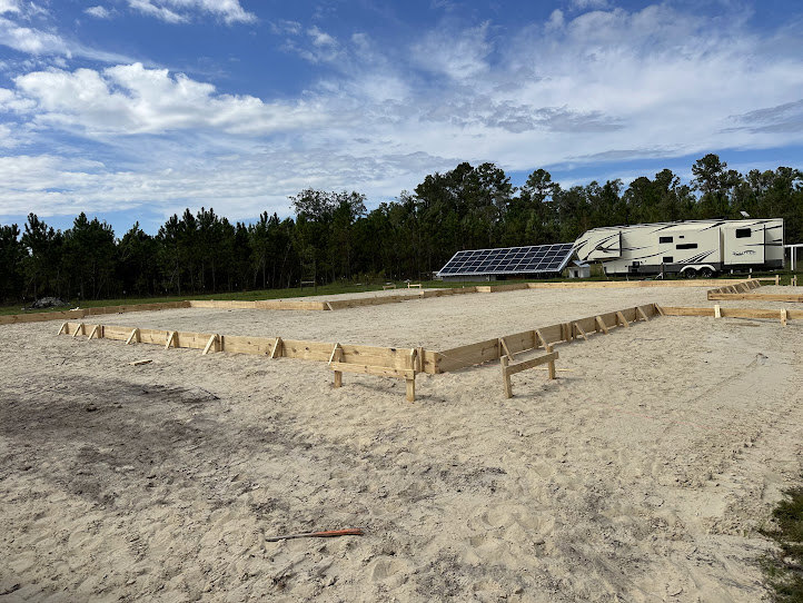 Wide sandy yard with a white camper featuring black stripes, surrounded by mature trees under a partly cloudy sky