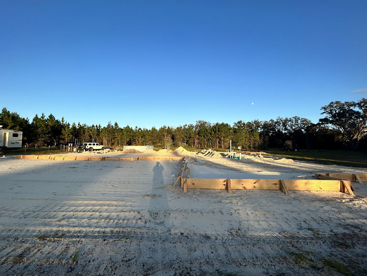 Framed construction site with bare ground, white trailer with window, shadow of person, leafless trees and blue sky in background