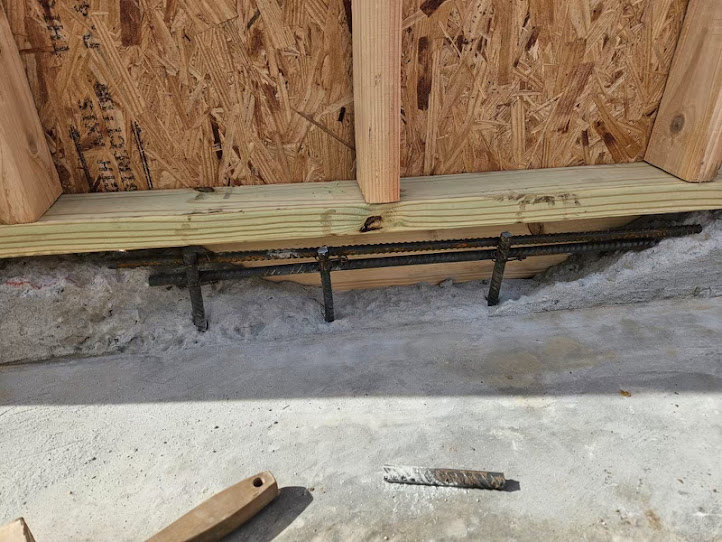 Wooden beam with exposed metal rods resting on outdoor plywood surface, surrounded by hardwood planks and lumber.