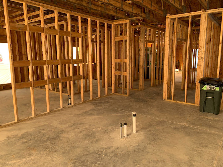 Wood-framed room under construction with exposed beams, concrete floor featuring white plumbing pipes, and a black trash can with a green label