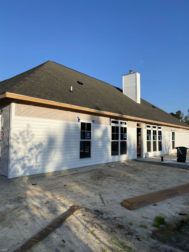 White siding house under construction with exposed windows, brown box on dirt ground, black trash can with white handle, unfinished roof against blue sky