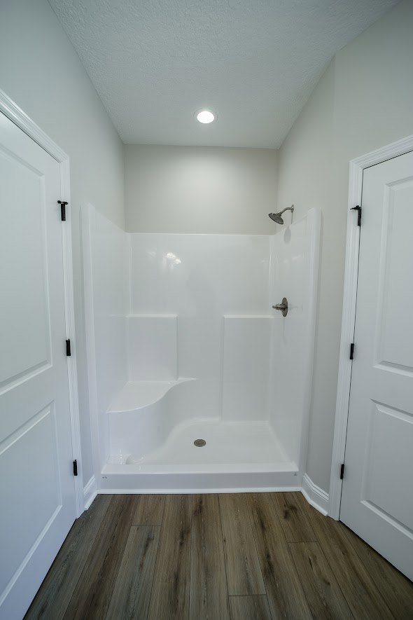 White shower with chrome shower head, wood floor featuring a black stripe, white trim, white door with black handle, white ceiling with recessed light.
