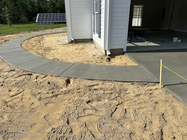 Concrete walkway bordered by soil and trees leading to the front entrance of a house with composite exterior materials