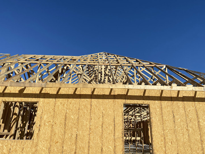 Wooden house frame under construction with exposed beams and plywood, set against a clear blue sky