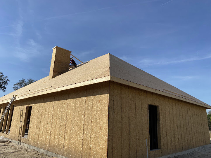 Person standing on shingled roof of two-story home with white siding, large windows, and surrounding trees under partly cloudy sky
