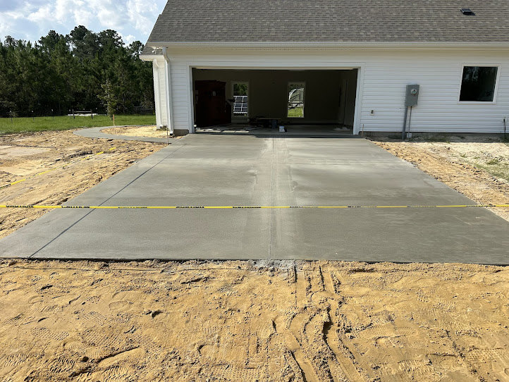 Concrete driveway leading to a garage with a window, grey roof with white trim, yellow and black caution tape on the ground, trees and sky in the background