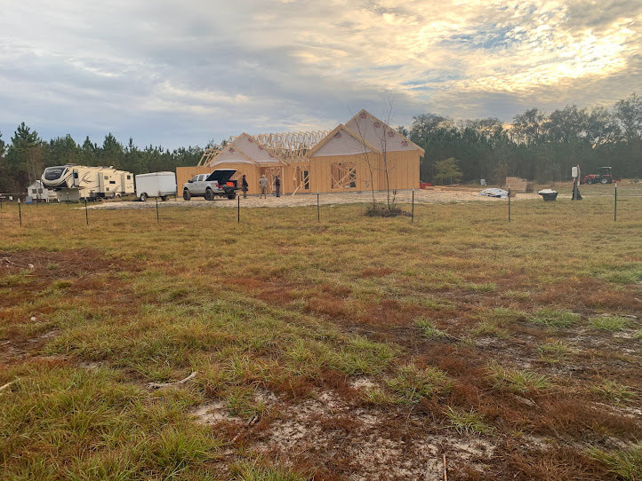 Wood-framed custom home under construction, surrounded by a chain-link fence and parked pickup truck, set on grassy lot with scattered trees and cloudy sky in rural landscape