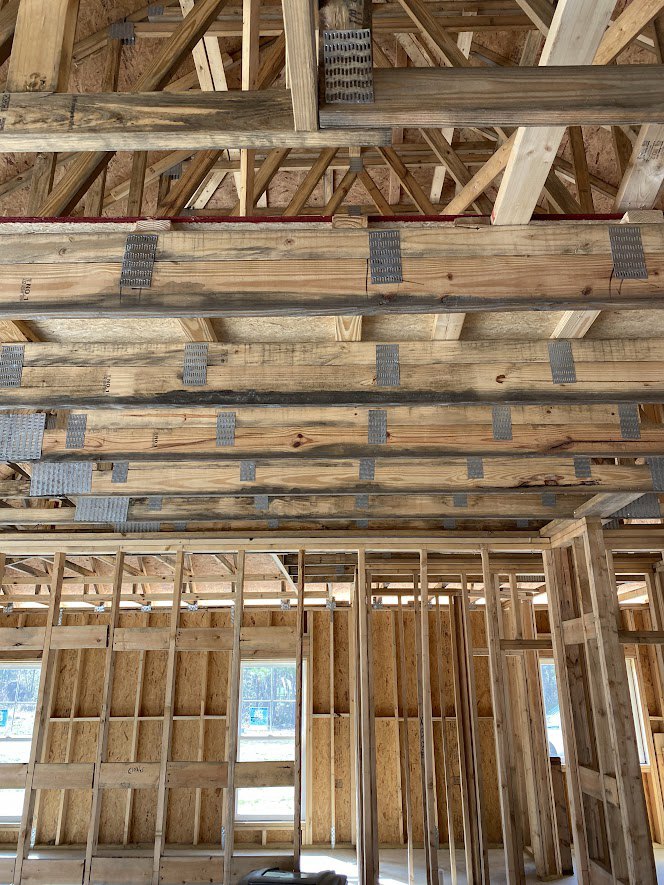 Exposed wood framing with beams, planks, and metal fasteners inside a partially constructed house; blue sign visible on window, insulation not yet installed