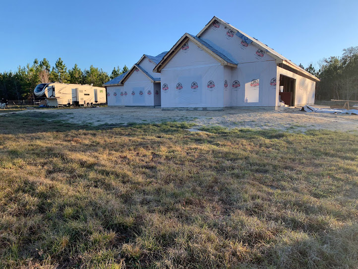 Partially built house with exposed framing and construction materials, white trailer parked nearby, grassy lot in foreground, clear blue sky overhead