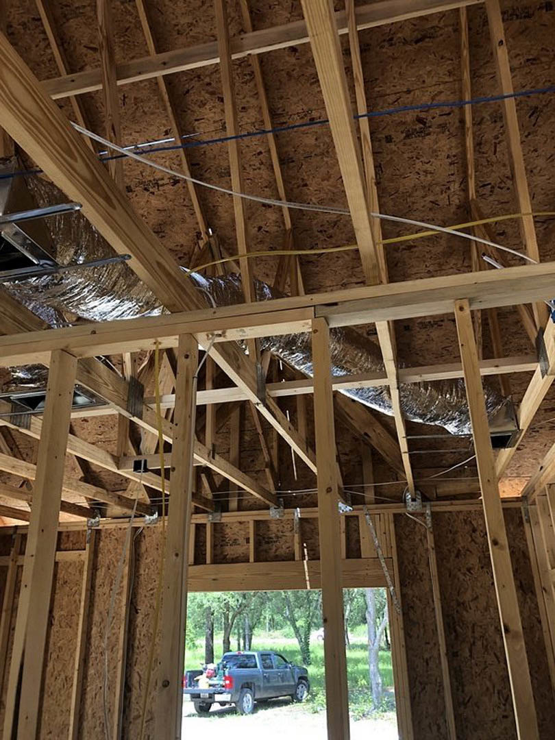 Wood-framed house under construction with exposed beams, metal pipes, and electrical wiring; grey truck parked nearby in a wooded area