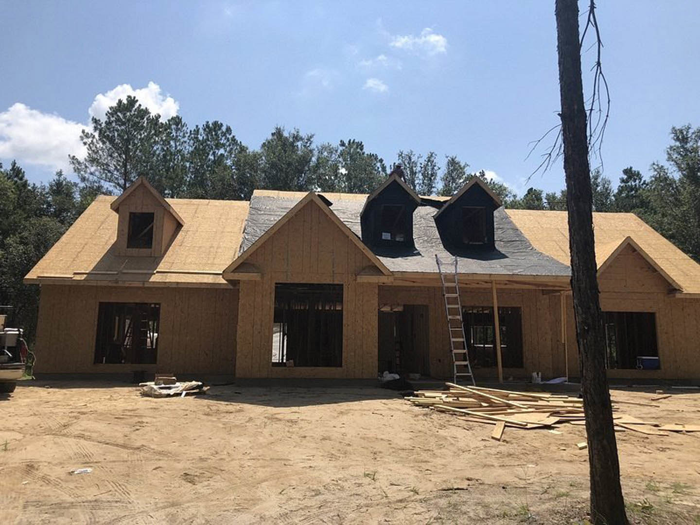 Framed house under construction with exposed wood, ladder leaning against unfinished wall, dirt ground scattered with wood pallets, large window opening, tree trunk in foreground