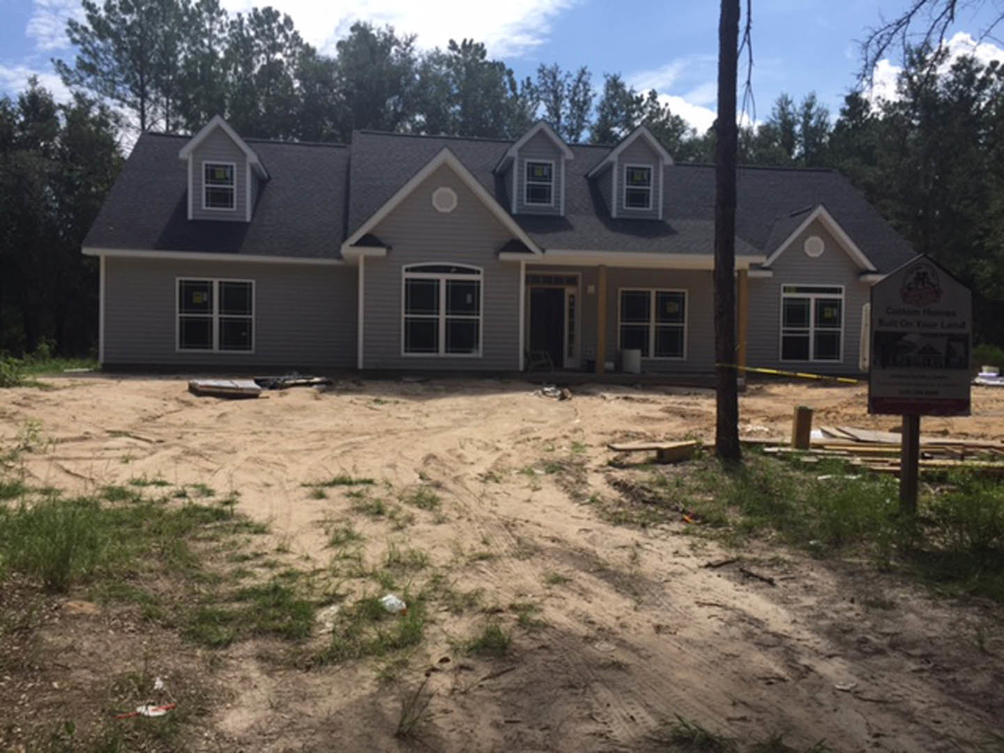 Wood-framed house under construction surrounded by trees, unfinished exterior walls, exposed windows, dirt yard with patches of grass, construction sign on a pole