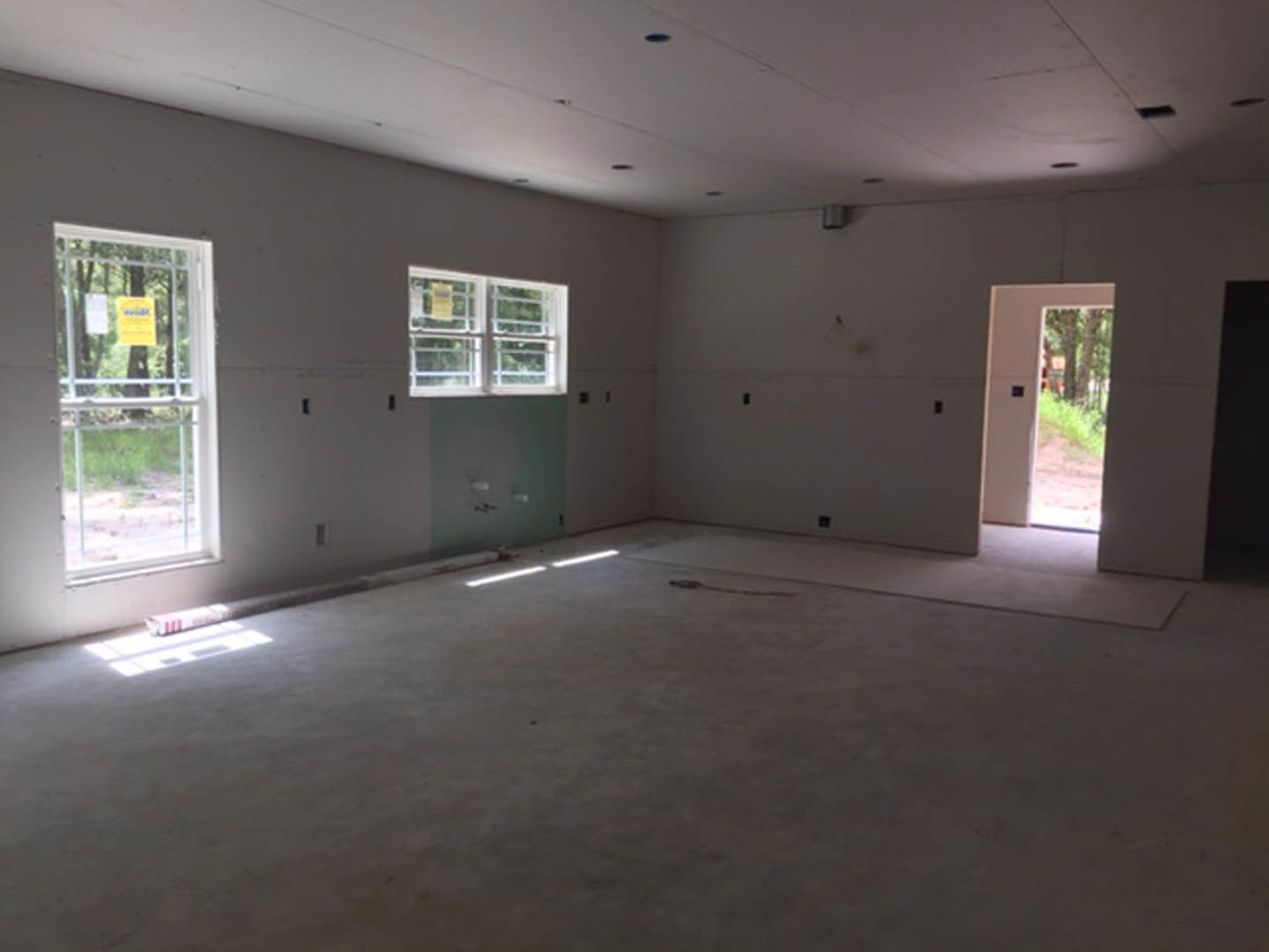 Modern interior room featuring white plaster walls, large windows with a blurry yellow sign, a white door with a black square detail, concrete flooring, and a rope lying on the