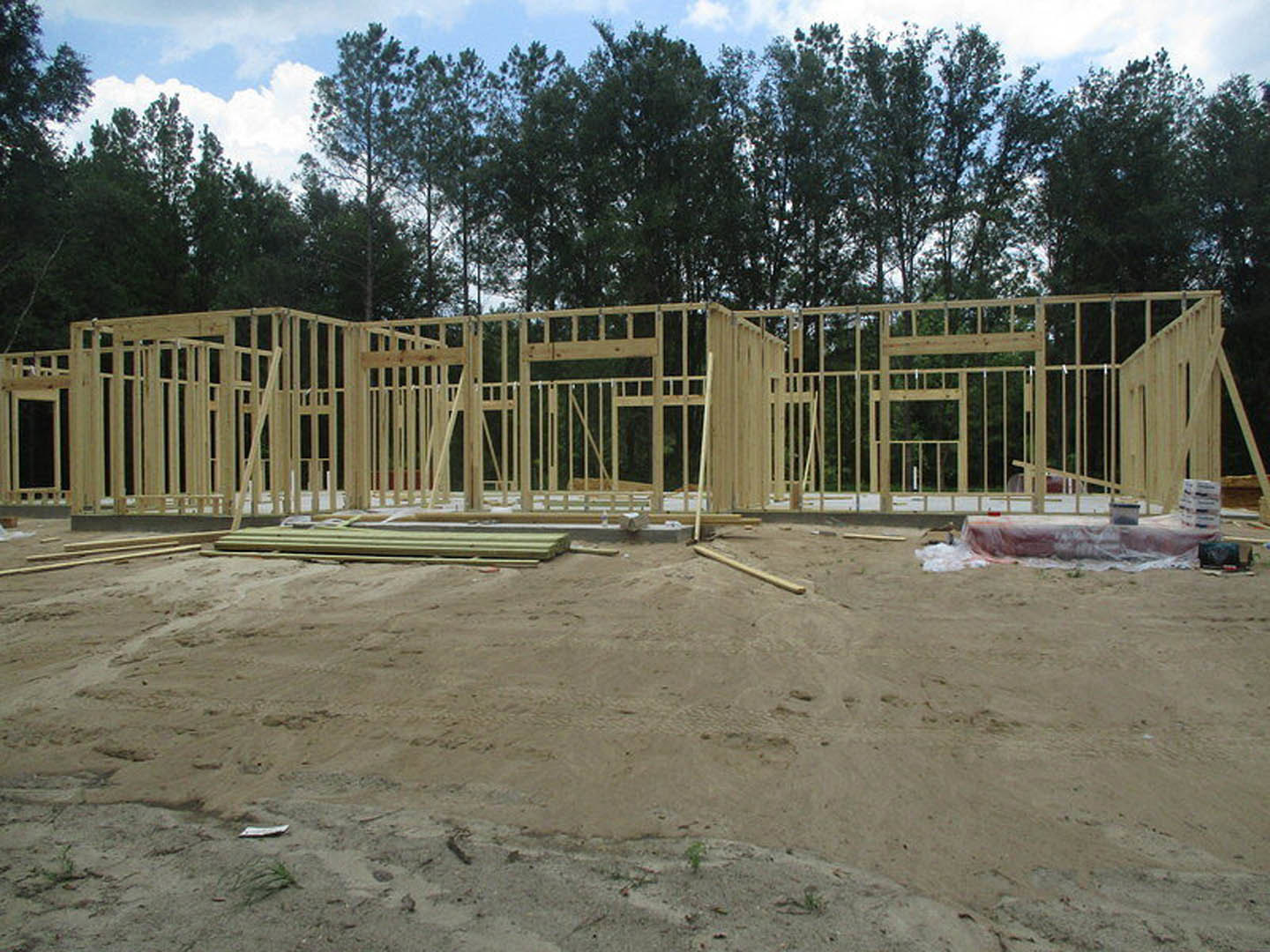 Wood-framed house under construction surrounded by tall trees, exposed beams and plywood, dirt ground, cloudy sky overhead