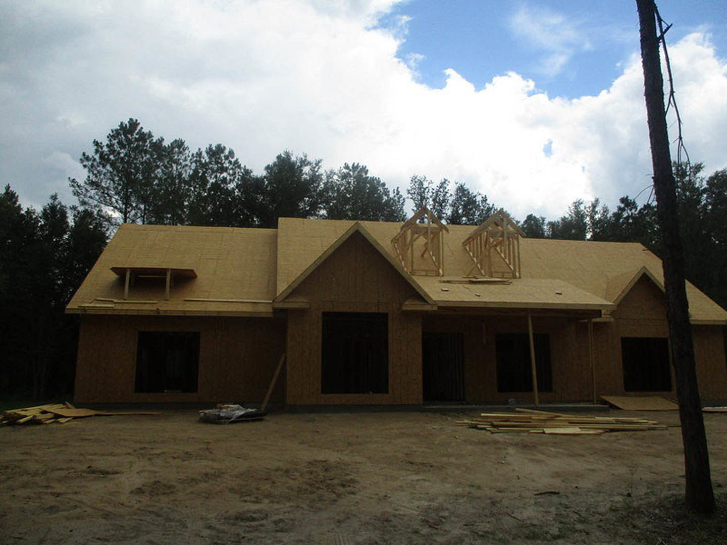 Wood-framed house under construction with exposed roof trusses, dirt ground, and mature trees in the background