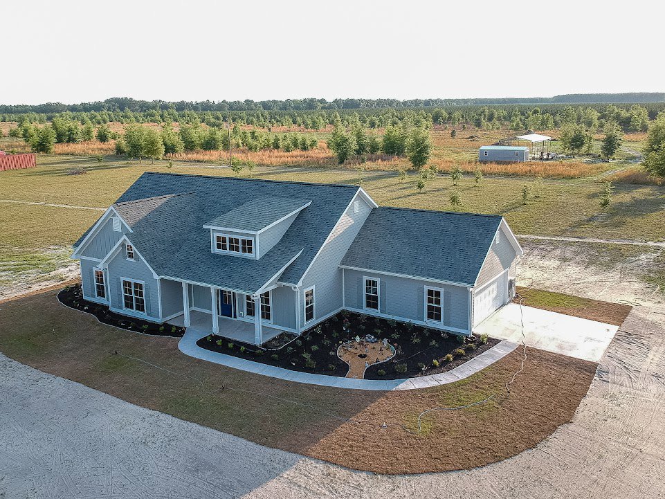 Two-story home with gray siding, large front lawn, landscaped garden beds with rocks and shrubs, multiple windows, and gabled roof with dormer window.