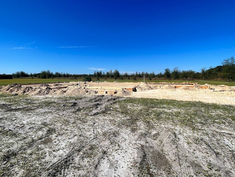 Dirt field bordered by green trees under a blue sky with scattered clouds, patches of grass and piles of sand visible in the foreground