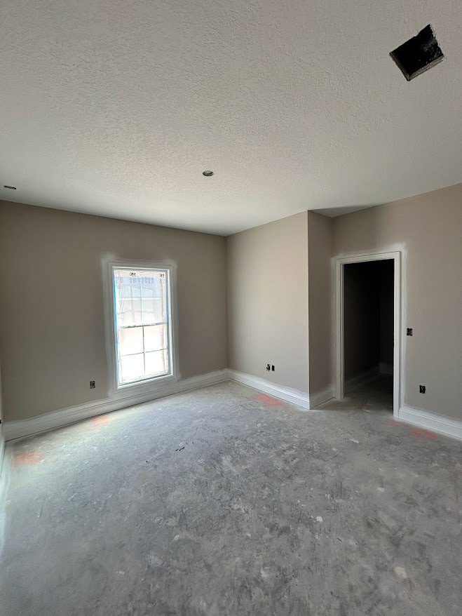 White plaster walls and ceiling with recessed spotlight, grey cracked flooring, window covered in plastic sheeting, white door with black hardware, black square object on white