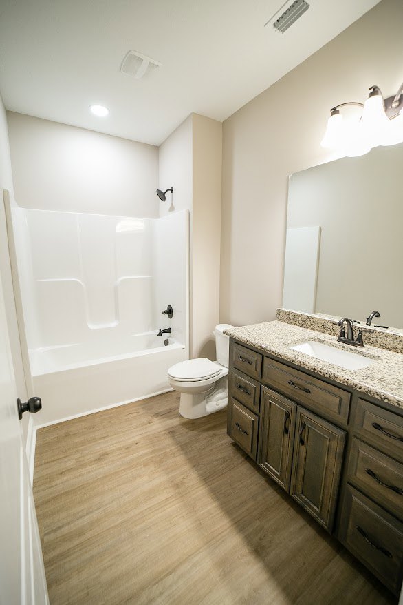 Bathroom with white toilet, rectangular sink on stone countertop, tile floor, wood cabinetry, and three-bulb wall light fixture above mirror