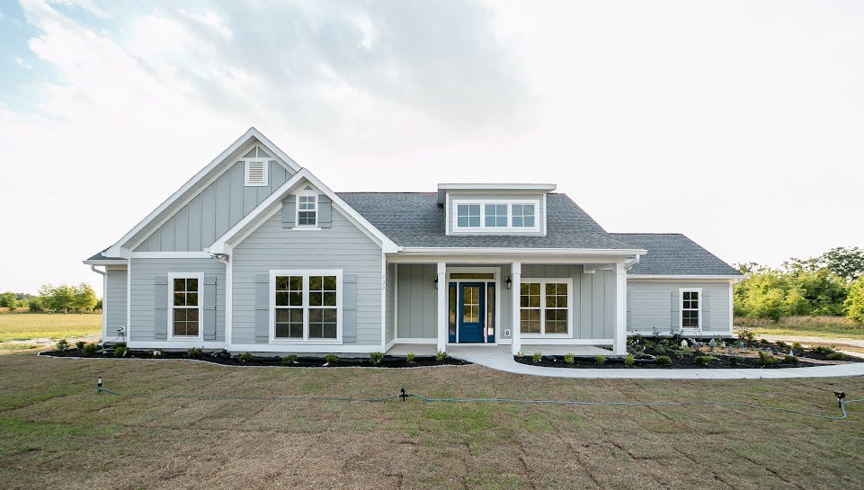 Two-story house with white siding, blue front door with white trim, multi-pane windows, green lawn with active sprinkler system