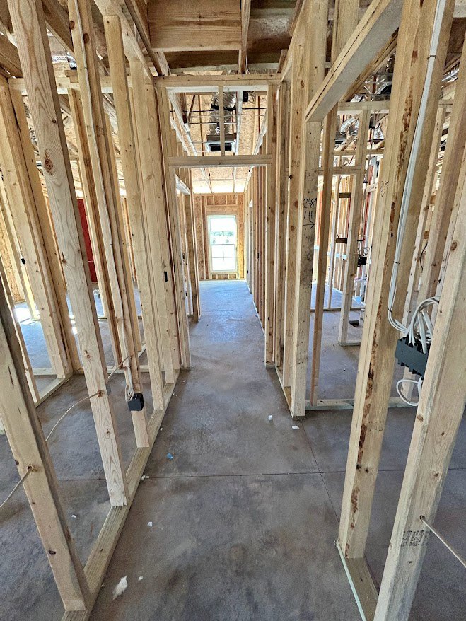 Exposed wood framing with vertical and horizontal beams, black electrical cord mounted to a wall, grey subfloor with a dark spot, unfinished interior construction with visible
