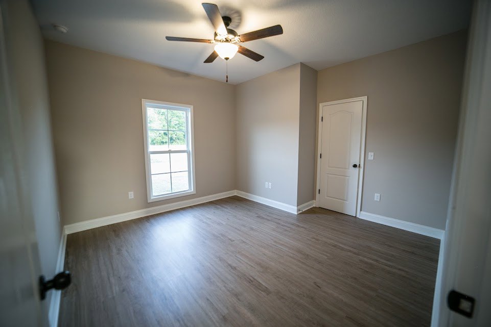 Ceiling fan with light fixture above hardwood floor, white walls, window showing trees, white door with black hardware