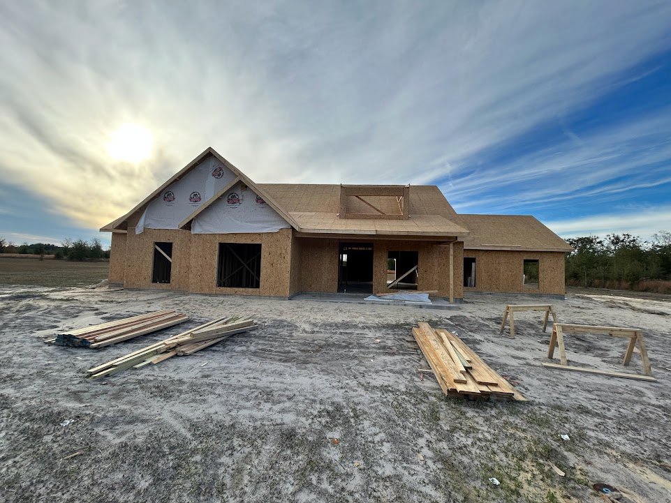 Wood-framed house under construction with exposed roof trusses, stacks of lumber on sandy ground, metal bars in doorway, cloudy sky overhead