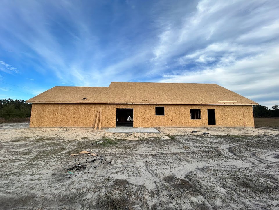 Partially built house with exposed framing, open doorway, dirt and grass foreground, pitched roof, blue sky with scattered clouds, trees lining the property