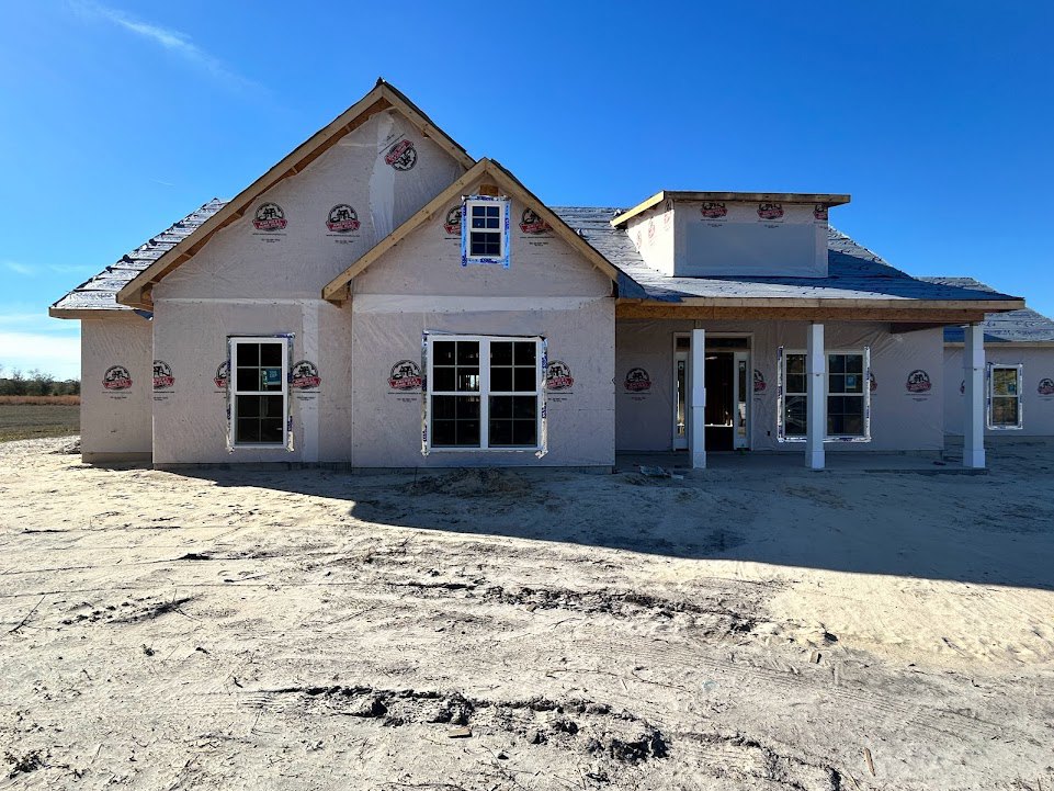 Framed custom home under construction with multi-pane windows, white door, dirt yard, white signpost, and blue sky with scattered clouds