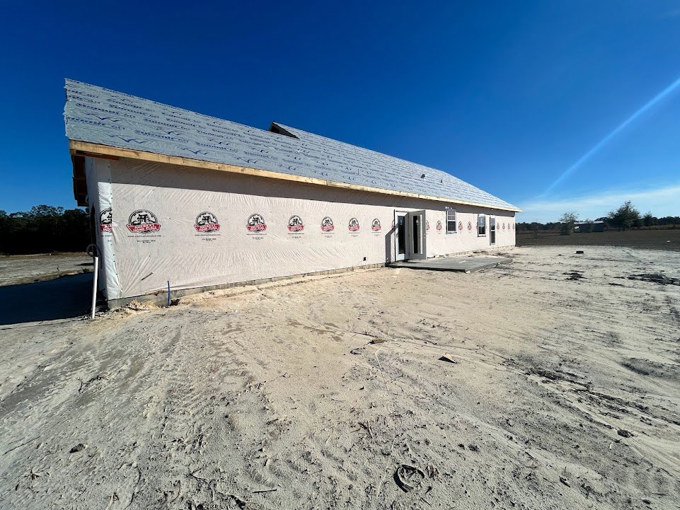 Partially built house with exposed framing, unfinished exterior walls, and construction materials scattered across a dirt field; blue sky with clouds and a faint rainbow overhead