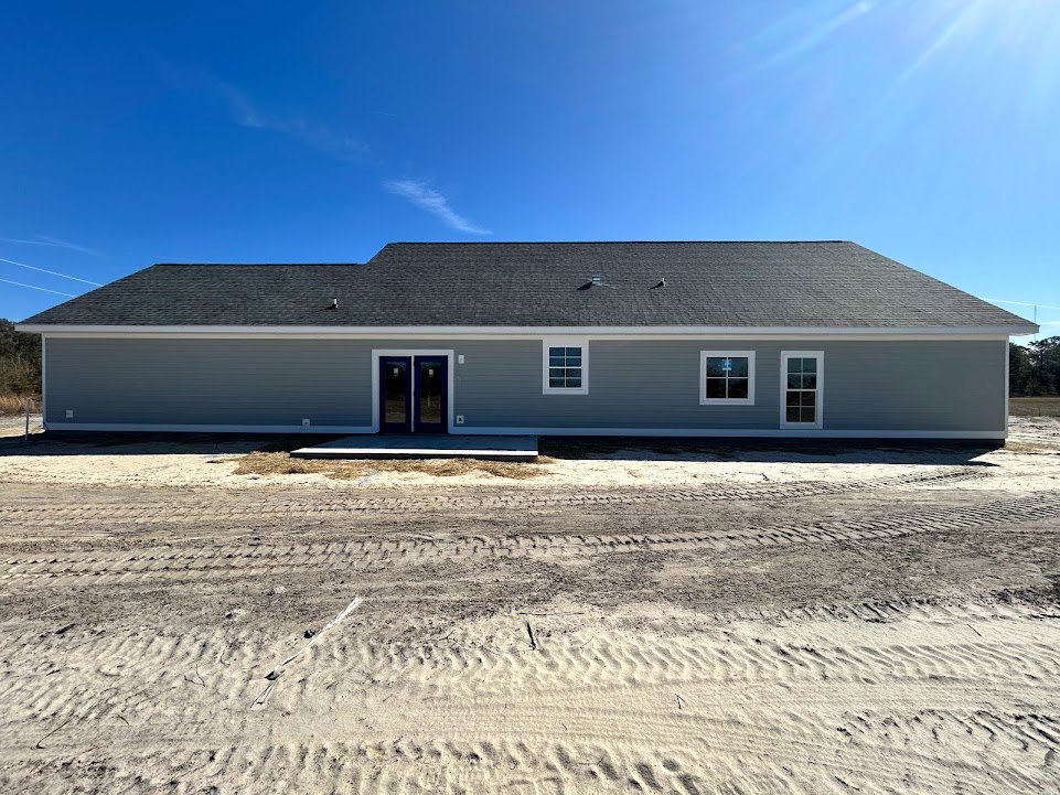Grey house with blue double doors featuring glass windows, tire tracks on sandy dirt road leading to entrance, window displaying a sign, blue sky with scattered clouds overhead