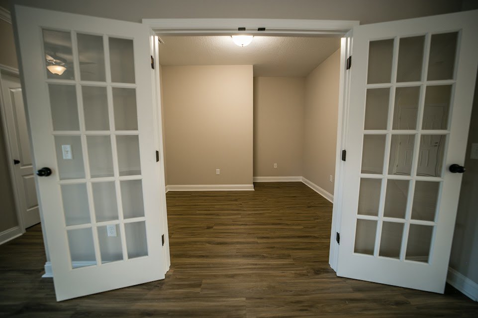 Room with double white doors featuring glass panels, wood flooring, white walls, and a visible electrical outlet