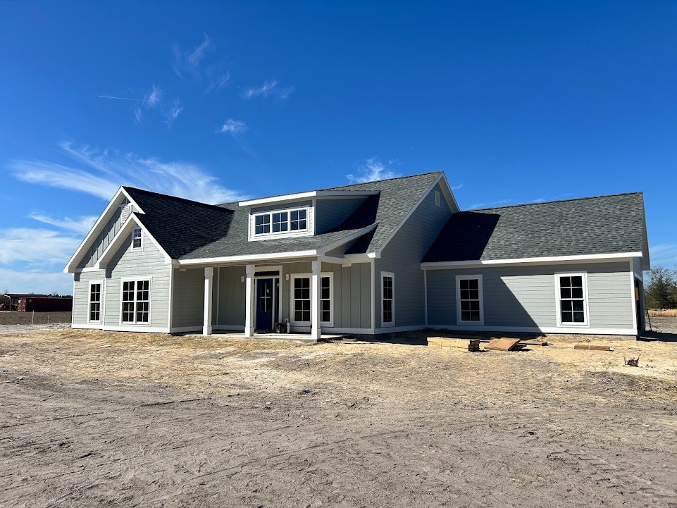 Partially built house with white-framed windows, exposed siding, and dirt lot under clear blue sky