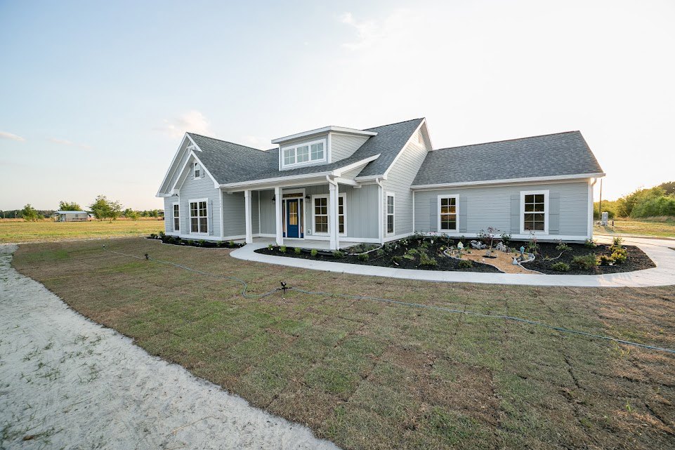 Two-story house with white siding, open front door, large windows, green lawn, garden hose on grass, dirt path leading to porch, cloudy sky overhead