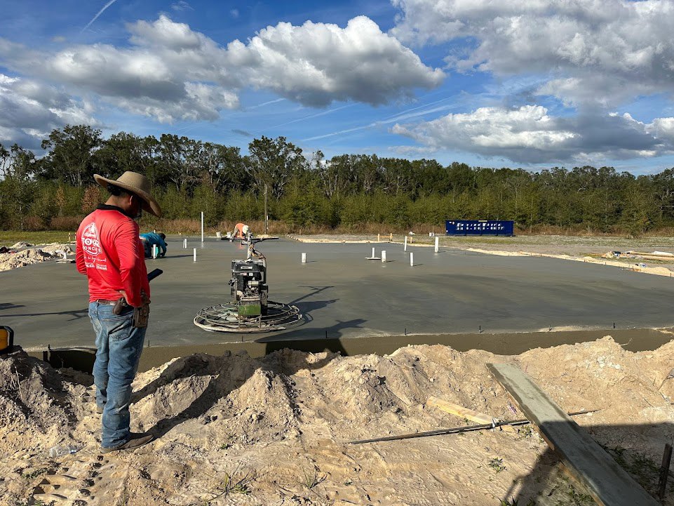 Man in red shirt standing on dirt near concrete slab, wooden beam lying on ground, construction equipment on slab, partly cloudy sky overhead, trees in background