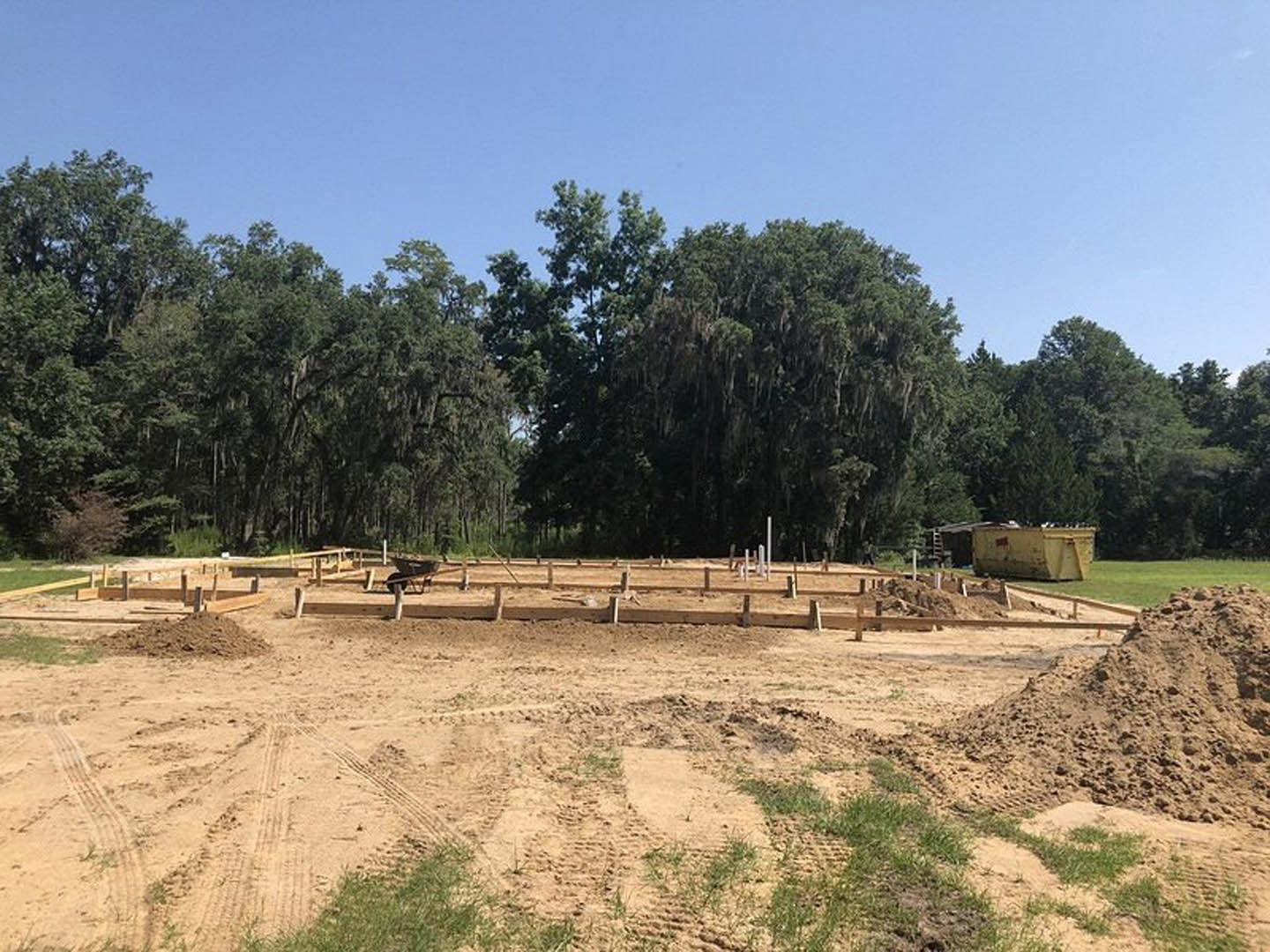 Dirt construction site with tire tracks, pile of soil, partially built wall, and moss-covered trees under blue sky in background