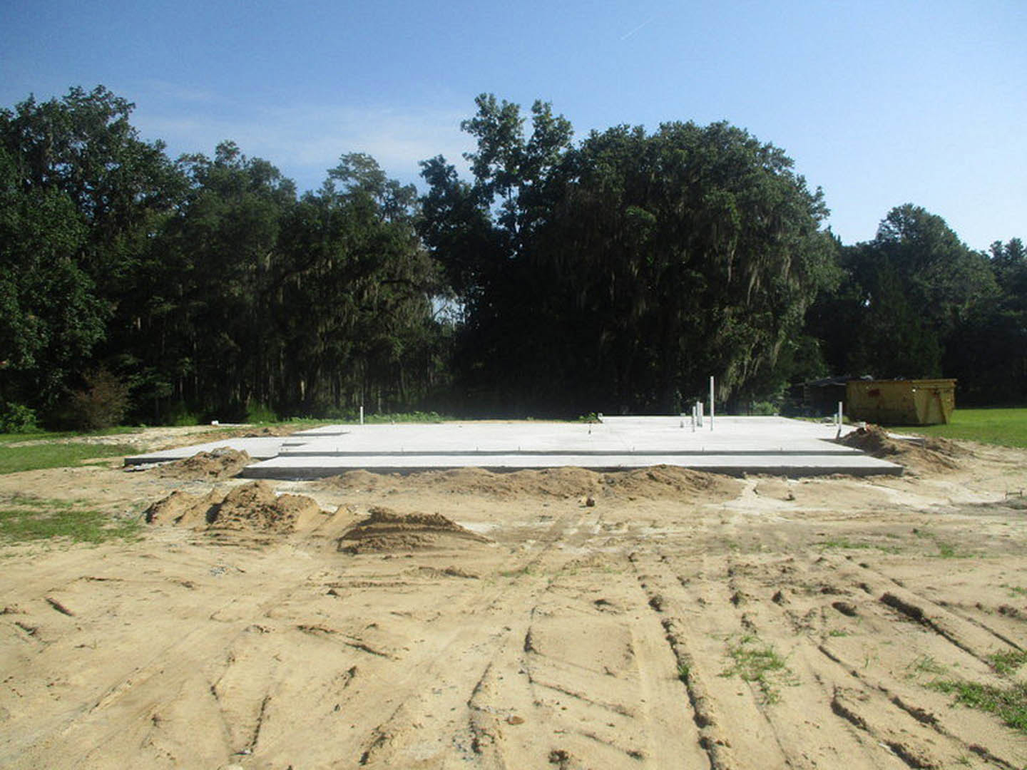 Dirt construction site with tire tracks, partially built wall, large leafy trees and blue sky in background
