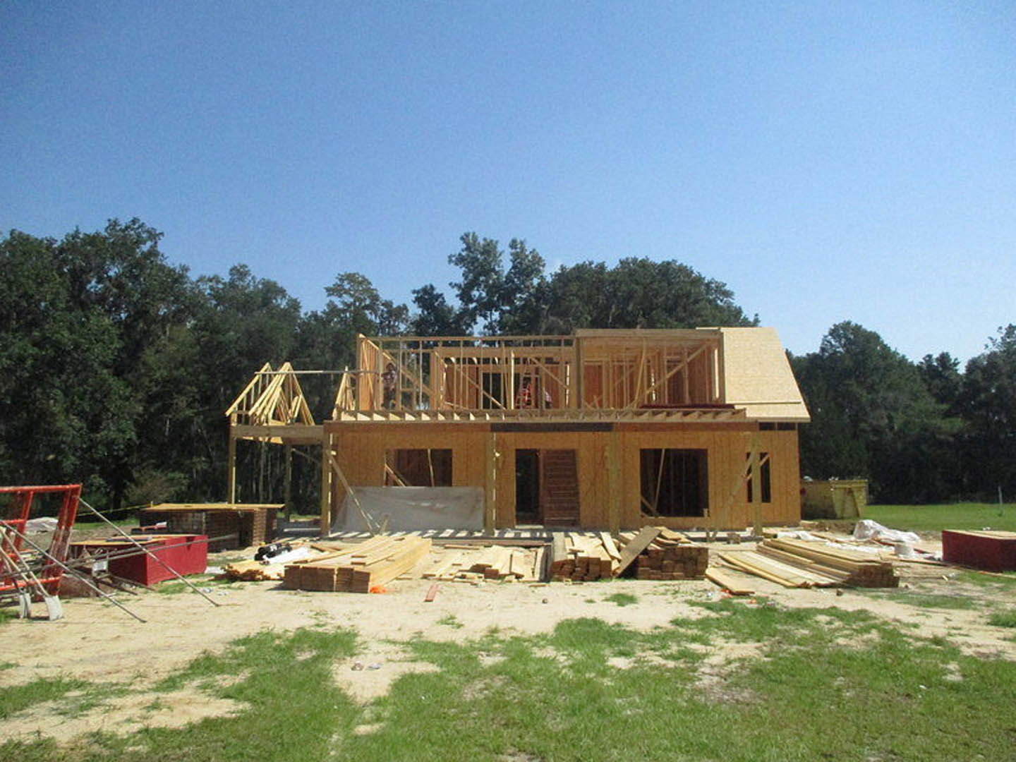 Framed house under construction with exposed wood beams, scaffolding, and stairs, surrounded by green grass and trees under a blue sky