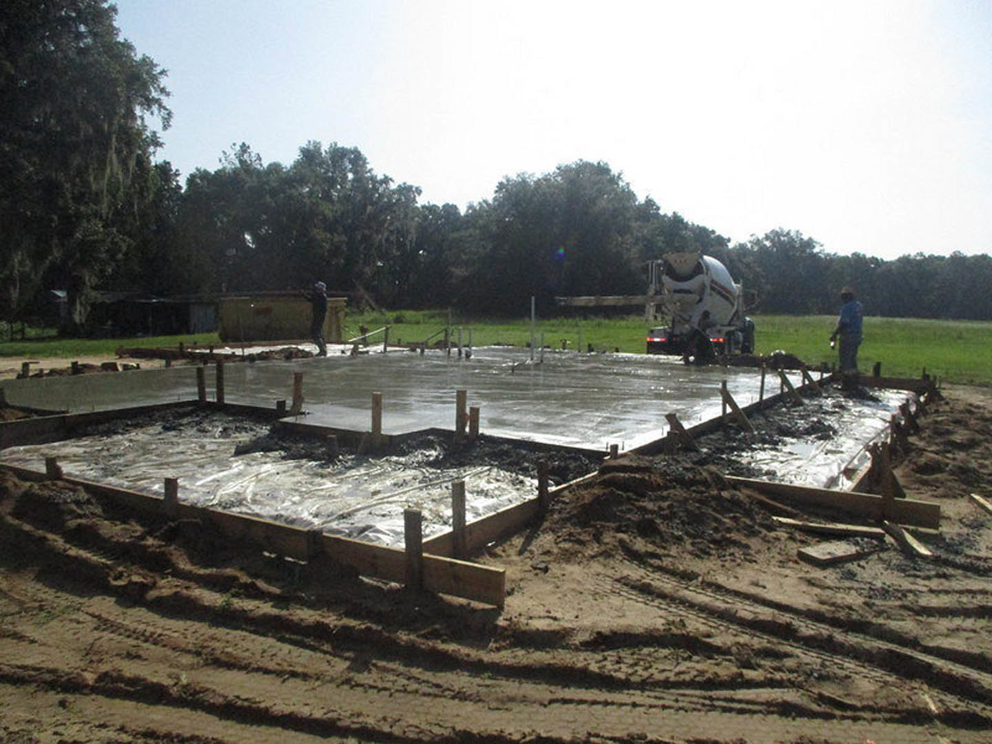 Freshly poured concrete slab foundation with wet surface, surrounded by muddy ground and patches of grass, concrete mixer truck parked nearby, wooden planks framing the edges