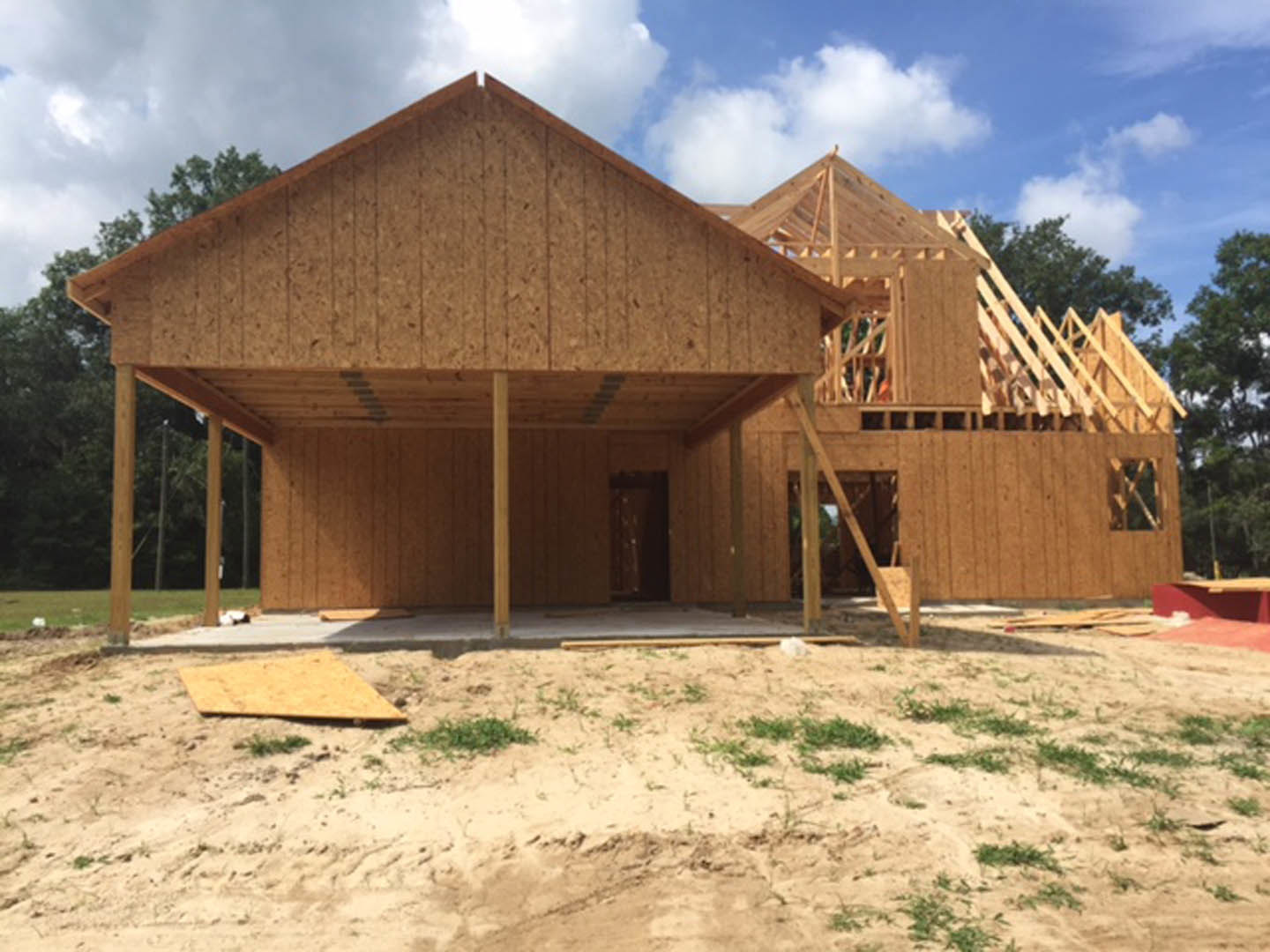 Wood-framed house under construction with exposed roof, dirt yard, scattered lumber, and small green bushes